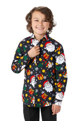 A smiling child is wearing a Suitmeister Santa Elves Black Christmas shirt, adorned with vibrant cartoon prints of Santa Claus, gifts, and stars. The child, who has medium-length curly hair, stands against a white background while holding one of the shirt's buttons.