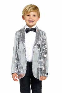 A young boy stands smiling, sporting a Suitmeister Sequins Silver - Sequin Blazer paired with a white shirt and black bow tie. His blonde hair complements his formal attire, completed with black pants.