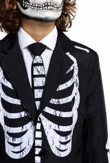 A person is dressed in the Suitmeister Skeleton Grunge Black, which includes a black suit adorned with a white skeleton ribcage print and a coordinating tie. Their mask, featuring a skull motif, conceals the lower part of their face, and they have curly hair.