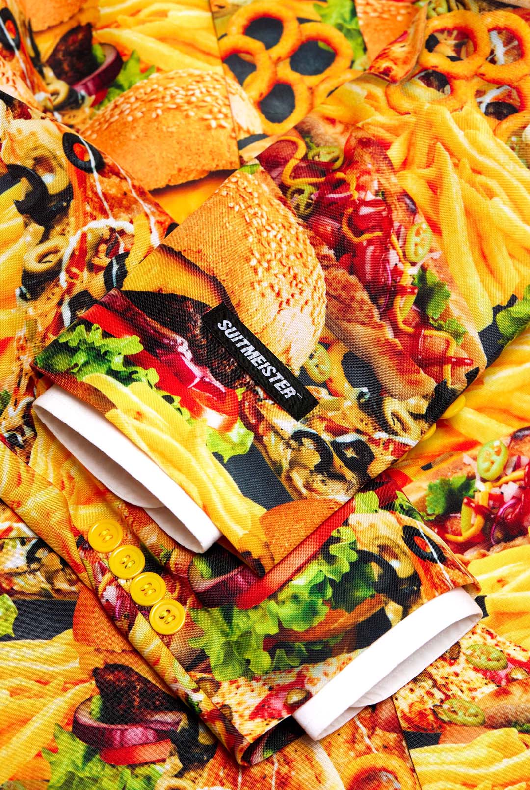 A close-up of the Snack Master by Suitmeister, a colorful suit jacket adorned with a vibrant food-themed print showcasing images of burgers, fries, pizza, and various other fast foods. The jacket is accented with yellow buttons and features a prominent black label displaying white text.