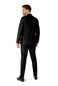 A man wearing the Solid Black suit from Suitmeister stands with his back to the camera, slightly turning his head to the left. The plain white background accentuates his formal attire.