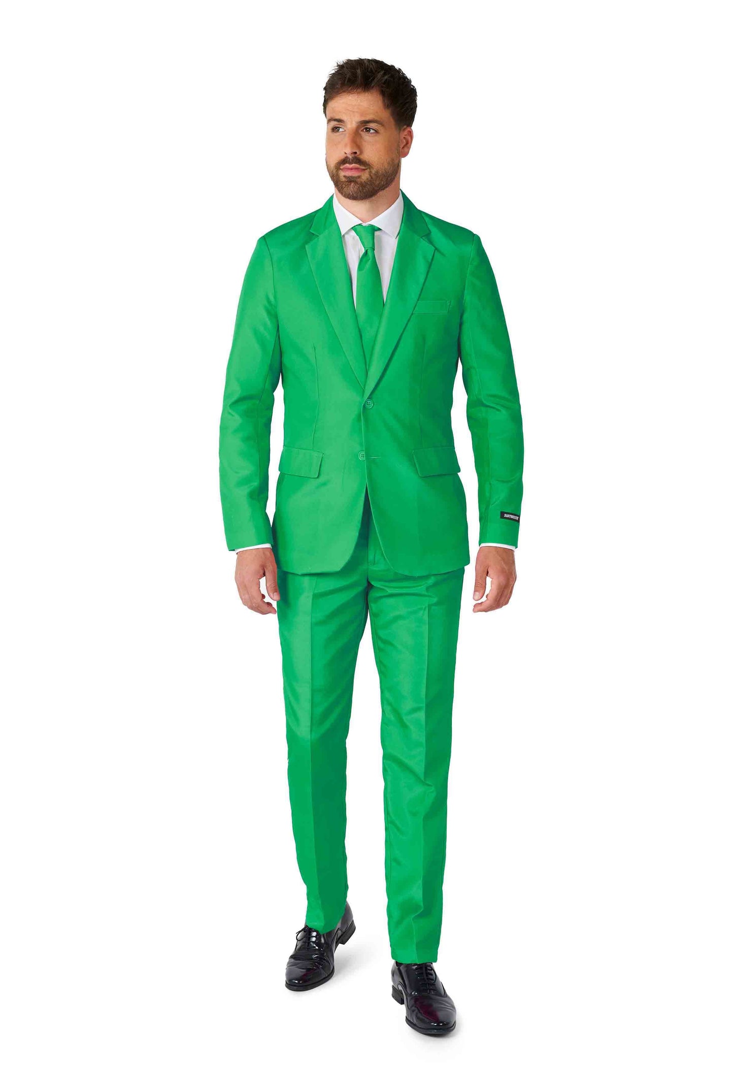 A man dressed in a Solid Green Suit by Suitmeister, paired with a white shirt and green tie, is standing against a plain white background. He has short hair and facial hair and is wearing black shoes.