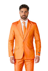 A man is standing, wearing the Suitmeister Solid Orange suit paired with a white shirt and an orange tie. He has short brown hair and a beard and is facing the camera. The suit features a single-breasted jacket with two buttons.