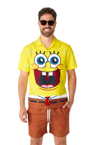 A man wearing sunglasses smiles while dressed in the SpongeBob outfit by Suitmeister, featuring a yellow shirt with a cartoon face that has big eyes and a wide grin, paired with brown shorts. The shirt resembles the well-known animated character, SpongeBob SquarePants.