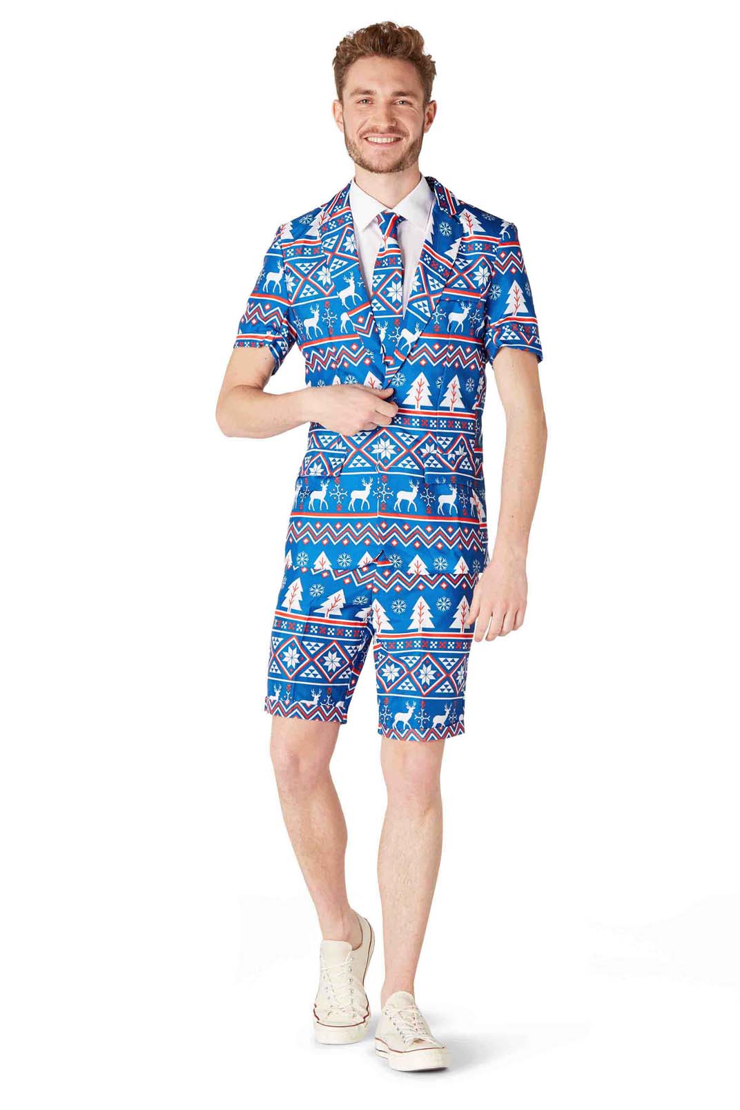 A person is dressed in the Suitmeister "Christmas Blue Nordic" ensemble, which boasts a festive holiday pattern adorned with reindeer and snowflakes. The outfit features a short-sleeved blazer, matching shorts, and a coordinating tie. They complete the look with white sneakers while smiling at the camera.