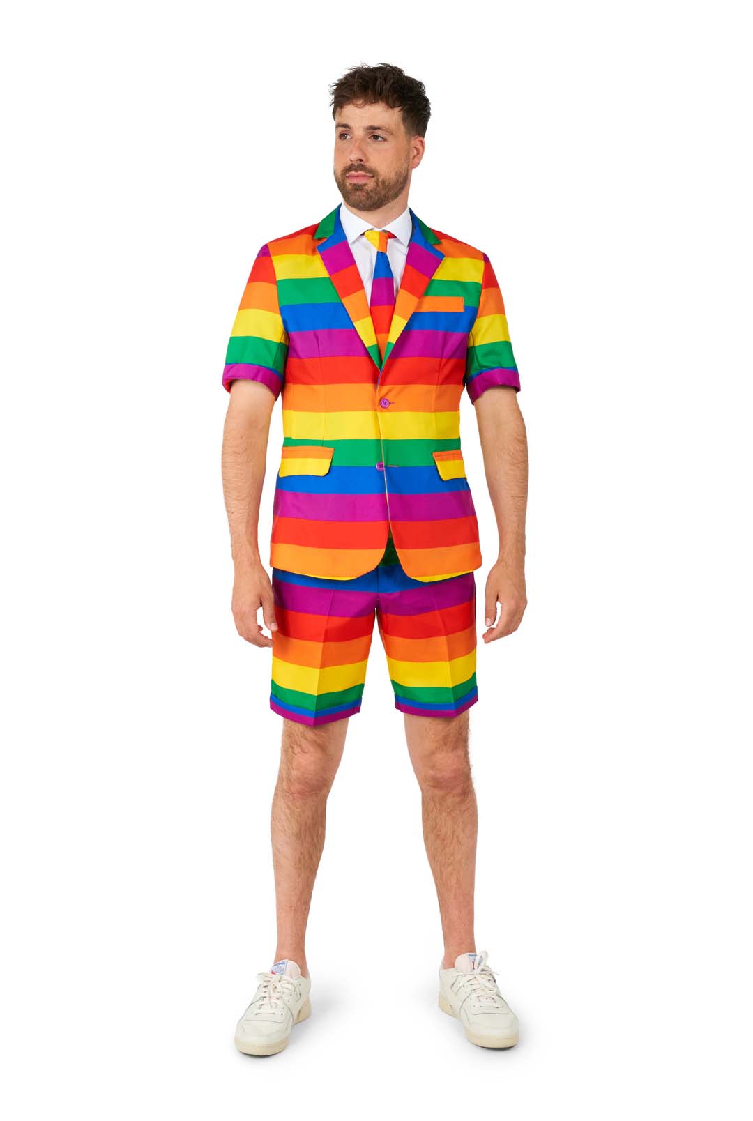 A man dressed in a Suitmeister Rainbow suit, featuring vibrant rainbow stripes with short sleeves and coordinating shorts. He pairs it with a white shirt, a rainbow tie, and finishes the look with white sneakers against a plain white background.