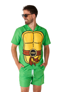 A man wearing sunglasses smiles while dressed in a Suitmeister TMNT outfit, featuring a green design with a cartoon shell print and a Teenage Mutant Ninja Turtles belt motif. The shirt and shorts set is reminiscent of a TMNT costume.