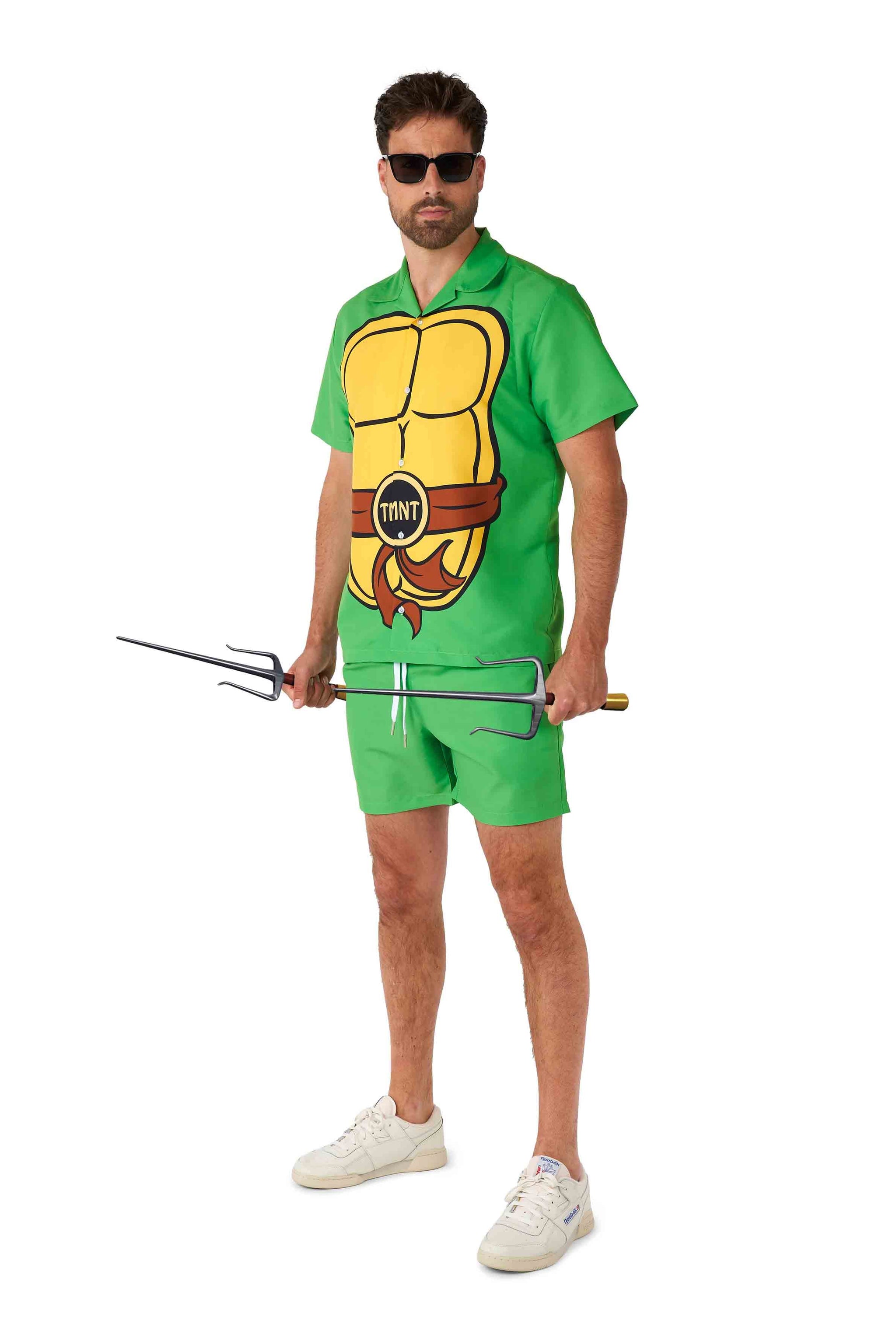 A man dressed in a Suitmeister TMNT outfit, complete with a green costume and holding two sai weapons, stands against a white background. He accessorizes with sunglasses, shorts, and white sneakers.