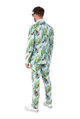 A person stands facing away wearing the Tropical Beers Blue - Festival suit by Suitmeister, adorned with a vibrant pattern of tropical leaves and fruits set against a light blue background. They complete the look with white sneakers and black sunglasses.