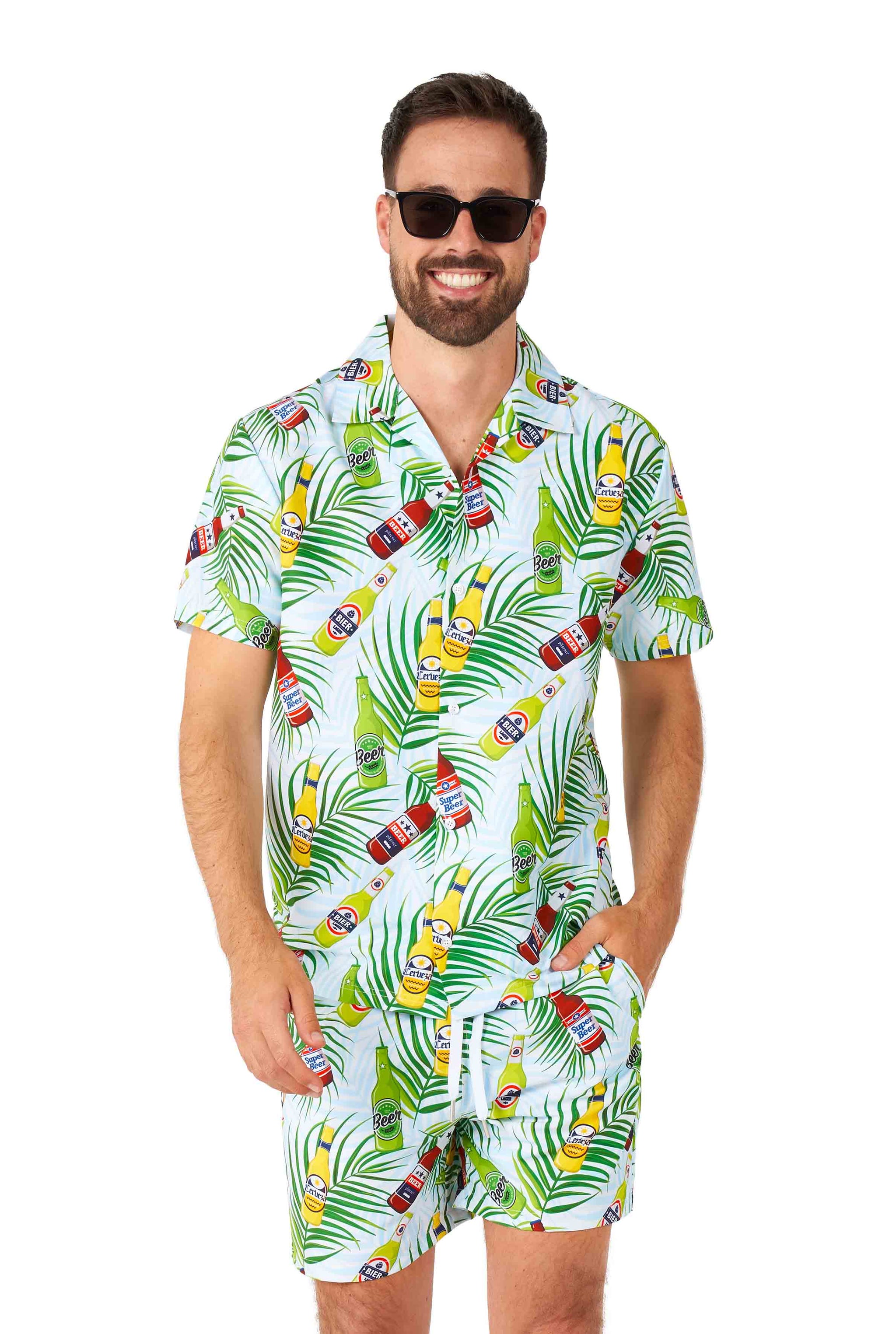 A man wearing sunglasses smiles while dressed in the Tropical Beers Blue - Festival suit by Suitmeister, featuring vibrant green leaves and bottle patterns. The shirt and shorts are matching, and his hands are in his pockets, set against a plain white background.