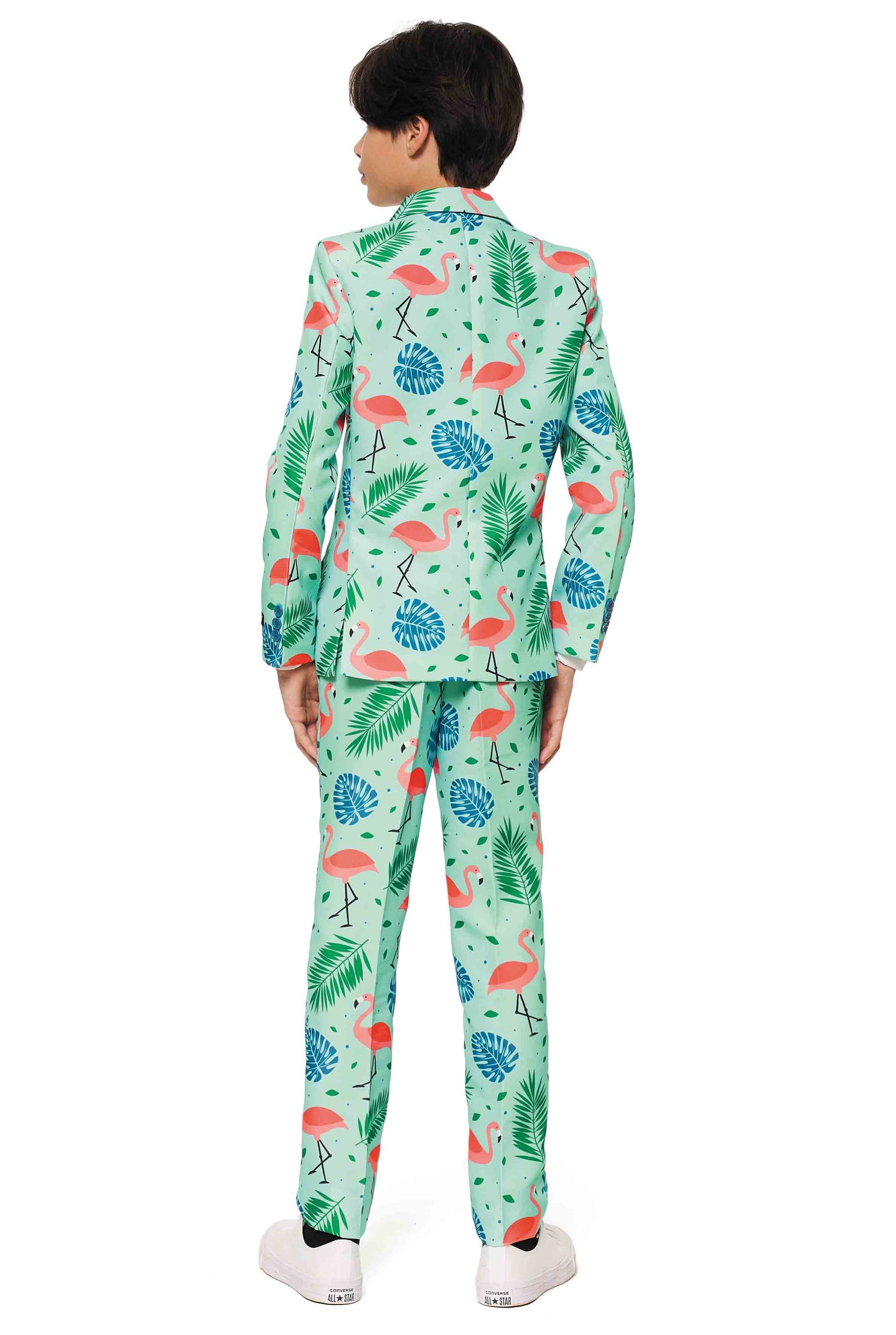 A young boy is wearing the Tropical suit from Suitmeister, featuring a playful pattern of pink flamingos and green tropical leaves on a light blue background. He completes the look with white shoes.