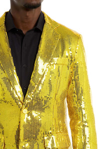 A person is wearing the Suitmeister Sequins Blazer in Yellow over a black shirt. The blazer is vibrant and reflective, catching the light with its sequins. The person’s face is not visible in the image.