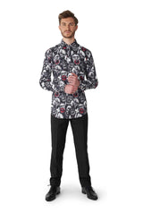 A man stands wearing the Zombie Characters - Zombie shirt by Suitmeister, paired with black pants and black shoes, against a plain white background. The shirt showcases a bold pattern of faces with red accents. He looks directly at the camera, hands clasped.