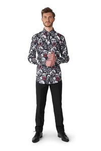 A man stands wearing the Zombie Characters - Zombie shirt by Suitmeister, paired with black pants and black shoes, against a plain white background. The shirt showcases a bold pattern of faces with red accents. He looks directly at the camera, hands clasped.